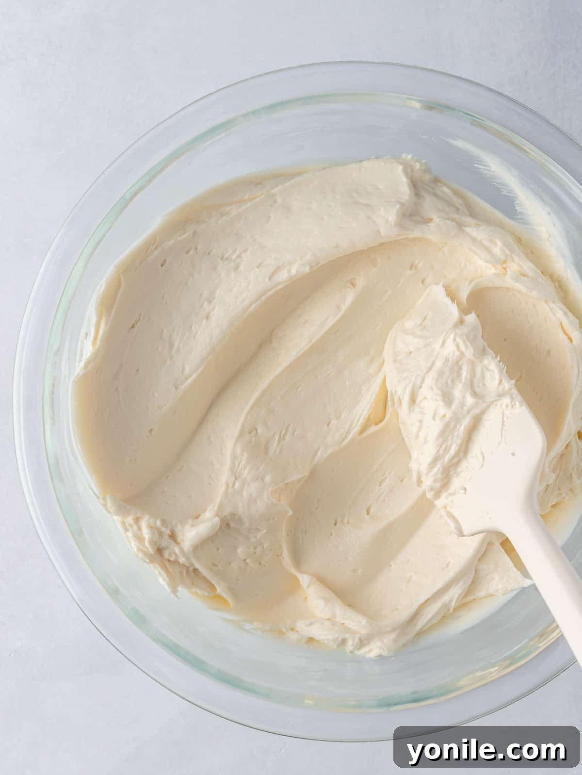 A bowl of creamy, white cream cheese frosting with a rubber spatula resting inside it.