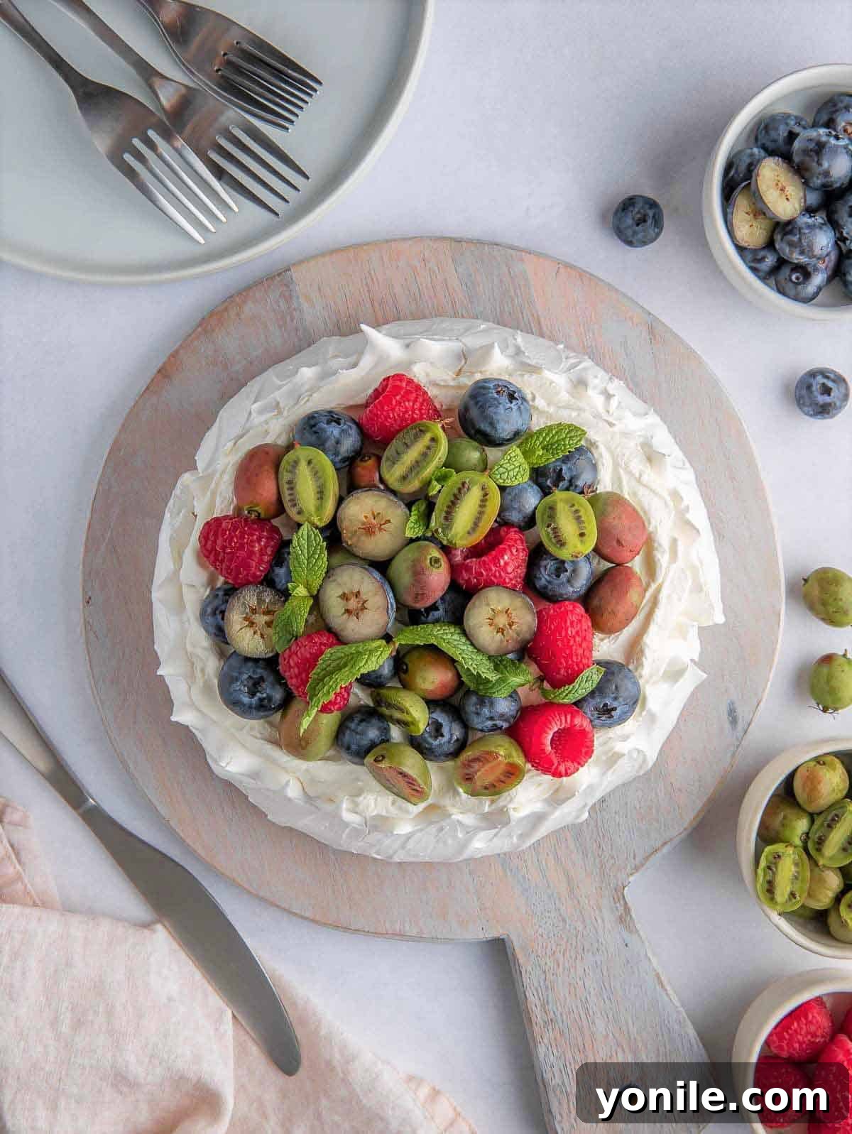 A top-down view of a fully assembled Berry Pavlova Cake, adorned with whipped cream and a generous assortment of fresh berries, resting on a rustic wooden board.
