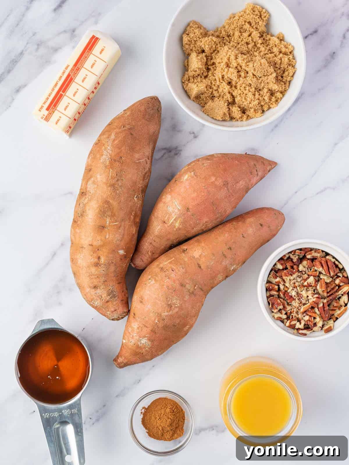 Fresh ingredients laid out for candied sweet potatoes, including whole sweet potatoes, butter, orange juice, maple syrup, brown sugar, cinnamon, and pecans