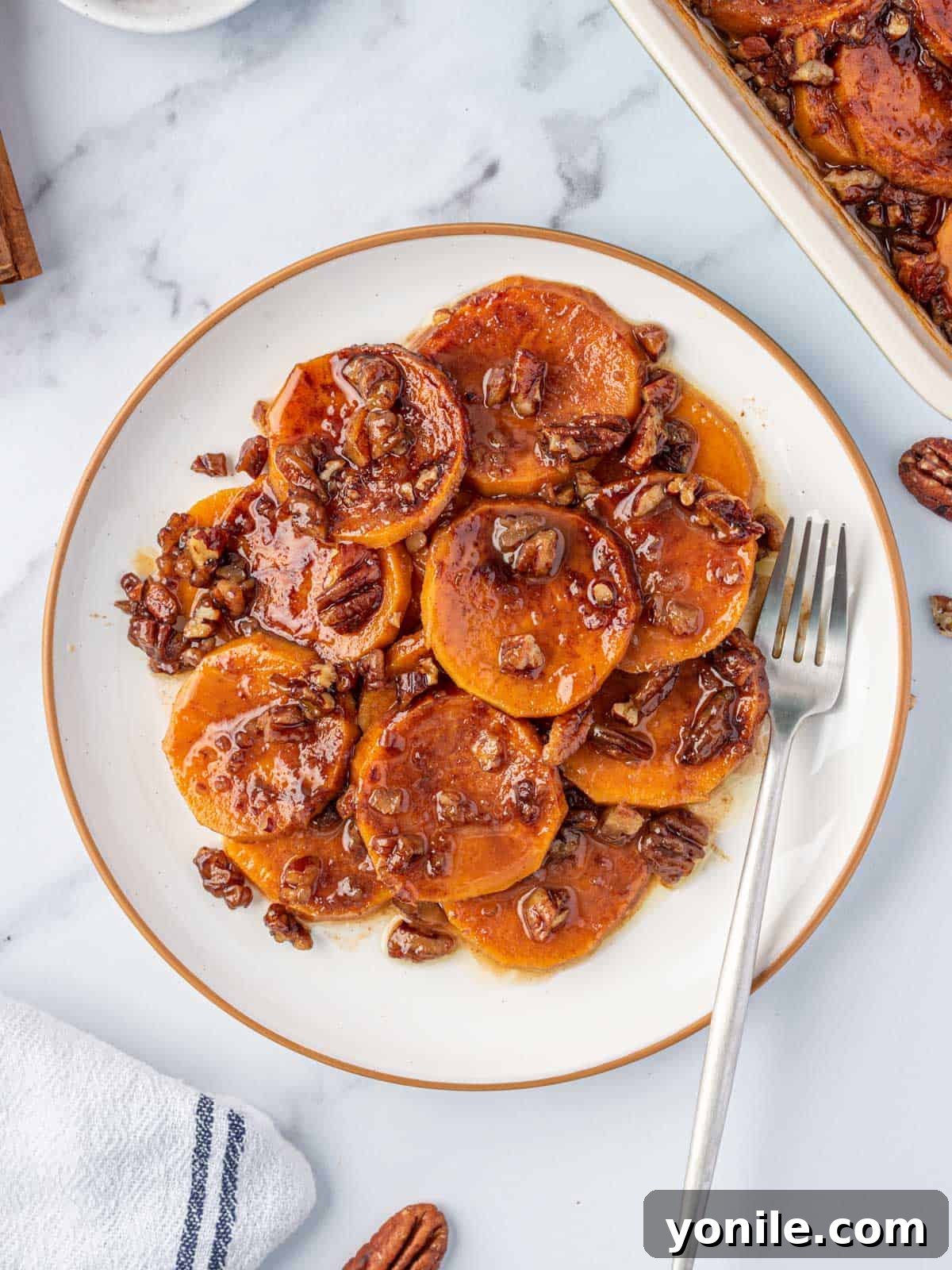 A serving of candied sweet potatoes on a white plate with a fork, showing the soft interior and pecan topping