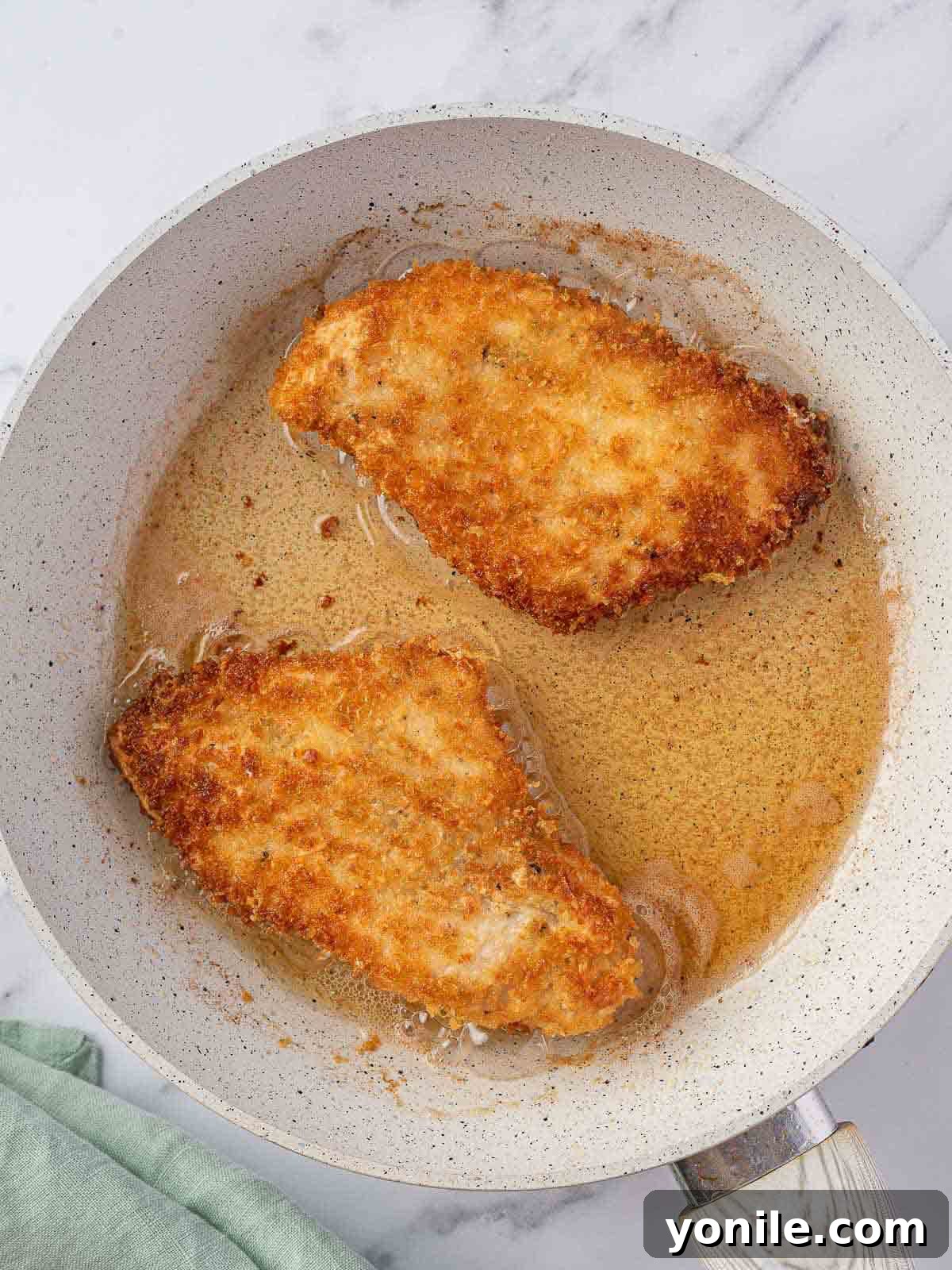 Two parmesan crusted chicken breasts pan-frying in a skillet until golden.