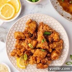 A white plate with Asian Lemon Chicken and a fresh lemon wedge. A bowl of fresh lemons is off to the side.