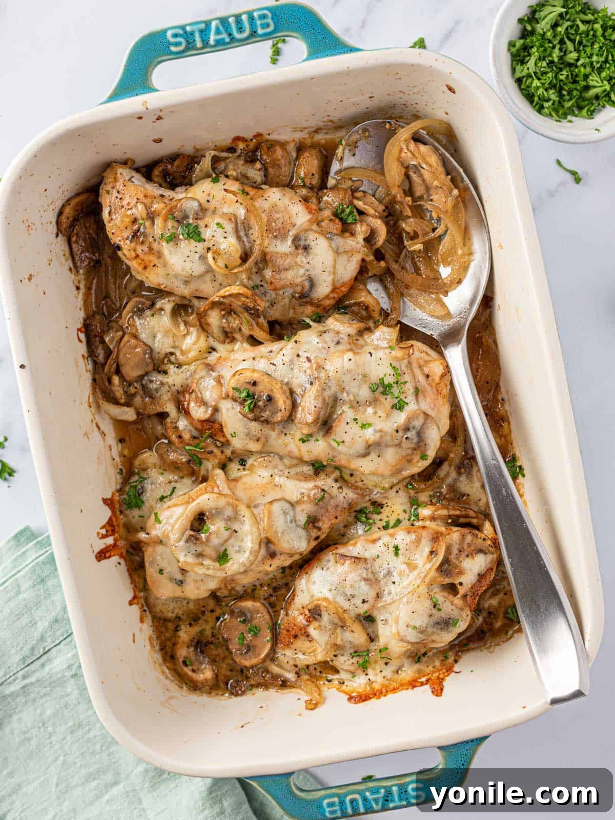A close-up of cheesy baked mushroom chicken in a baking dish with a serving spoon, highlighting the melted cheese.