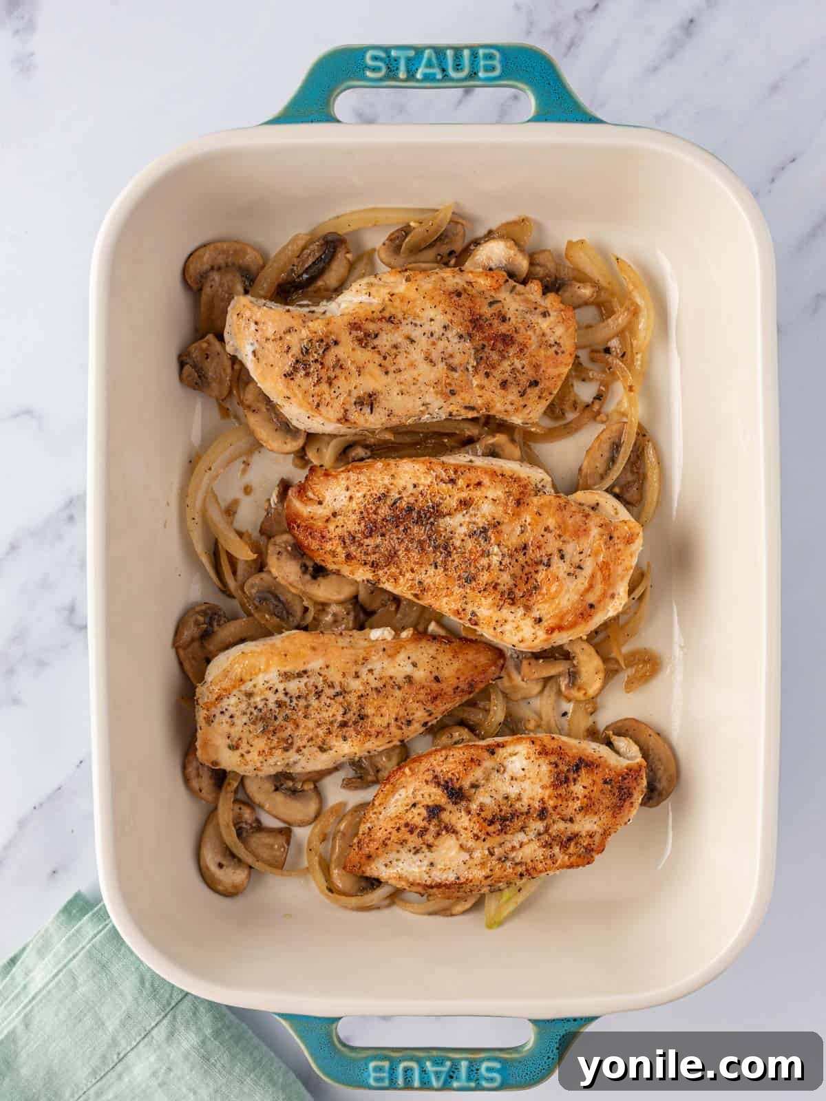 Half of the sautéed mushroom mixture spread evenly in the bottom of a blue baking dish, topped with seared chicken breasts.