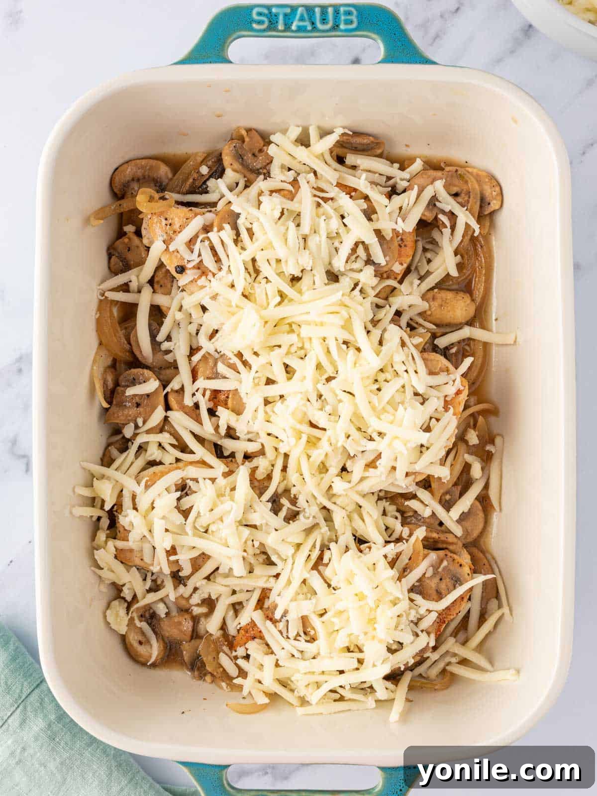 Shredded mozzarella cheese generously sprinkled over the chicken and mushroom layers in the baking dish, ready for the oven.