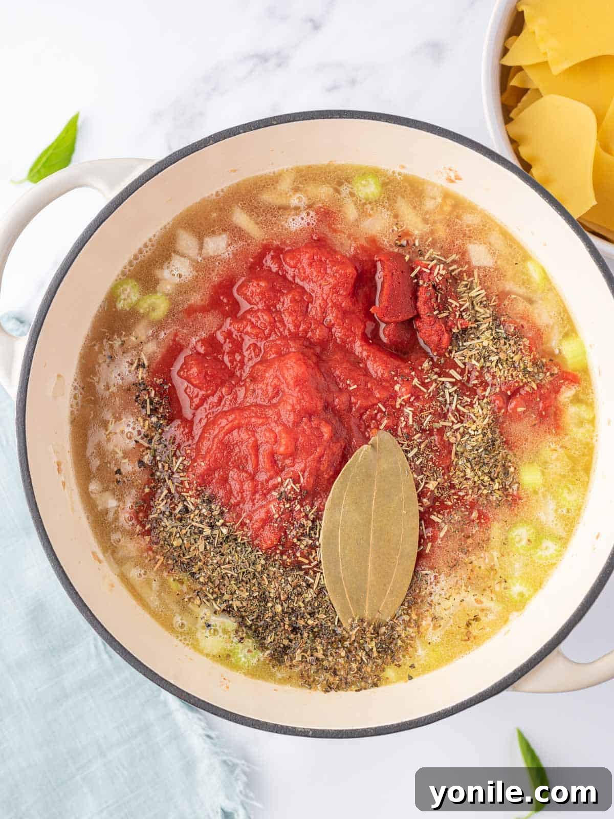 Process photo for making the broth with crushed tomatoes, herbs and spices and a bay leaf being stirred into the pot.