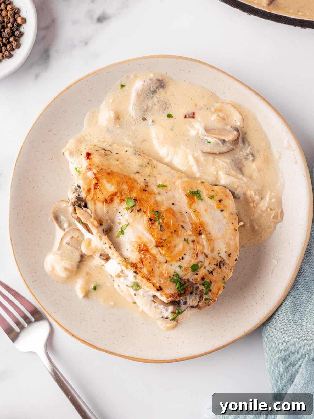 A plate with a mushroom stuffed chicken breast resting on top of the creamy garlic butter sauce. A fork sits to the left.
