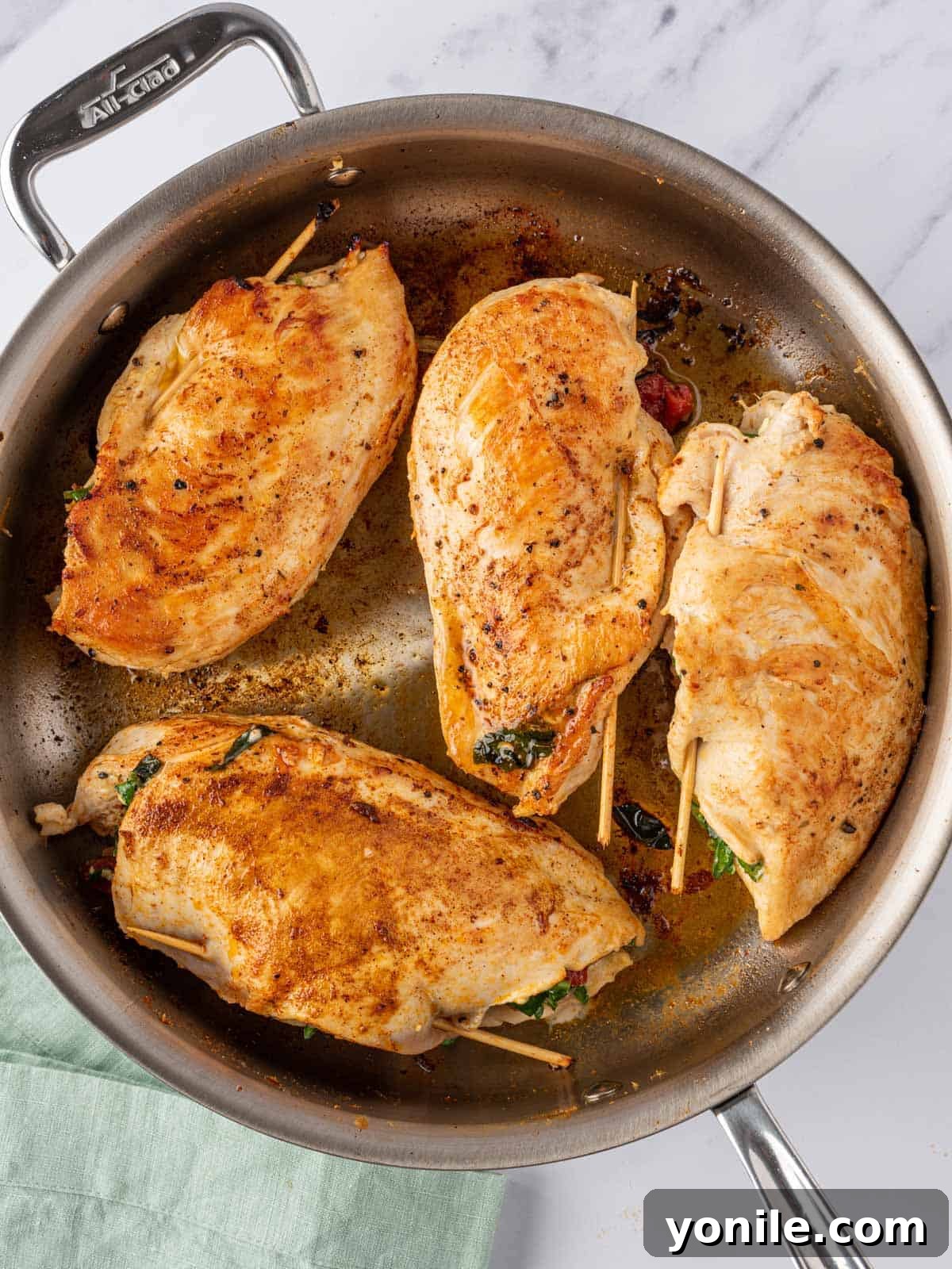Mediterranean stuffed chicken breasts being seared to a golden finish in a large pan on the stovetop.