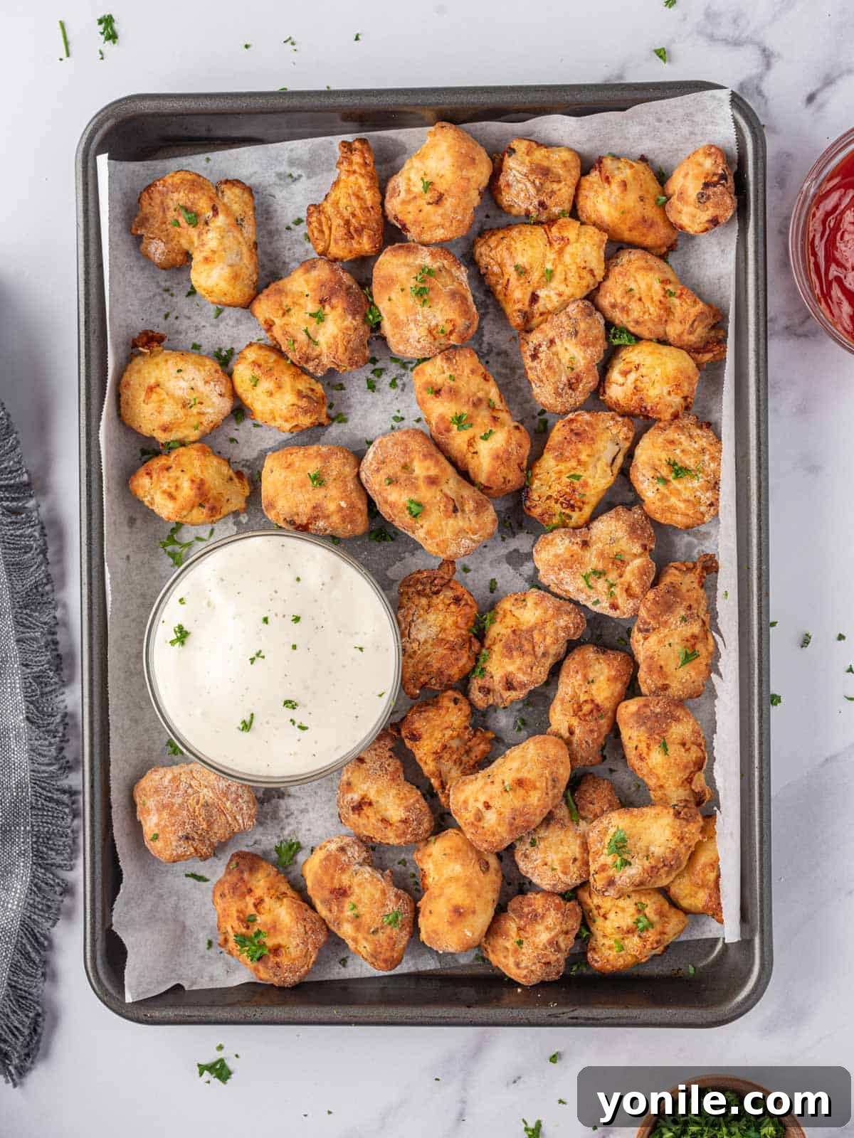 Golden Air Fryer Chicken Bites 2 Air fryer chicken nuggets on a parchment lined tray with a bowl of dip.