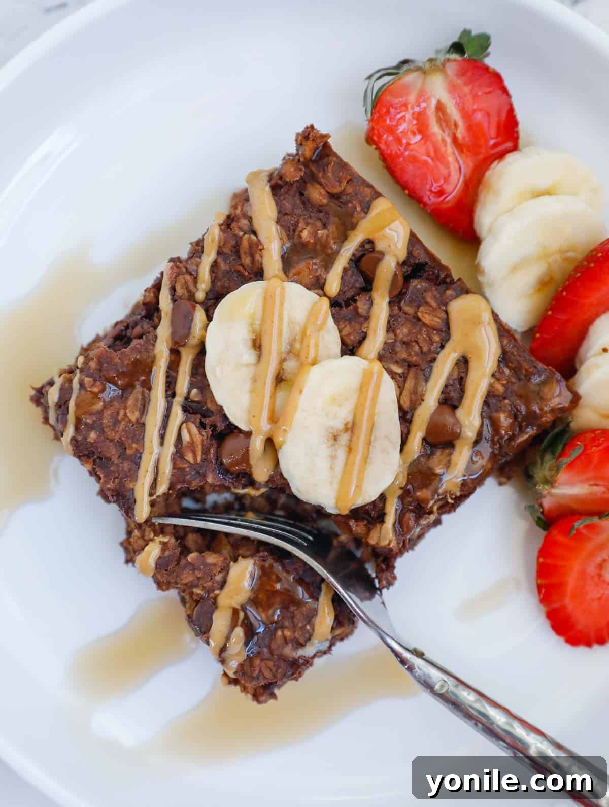 A delectable piece of baked chocolate oatmeal on a pristine white plate, garnished with freshly sliced bananas and a luscious drizzle of peanut butter, complemented by a touch of golden maple syrup.