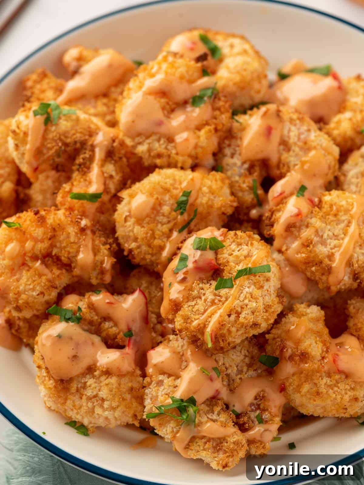 A close-up of a plate piled high with golden, crunchy air fryer panko shrimp, generously coated in a creamy, sweet, and spicy Bang Bang sauce.
