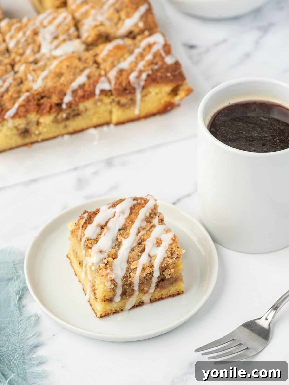 A perfectly sliced piece of keto coffee cake on a pristine white plate, accompanied by a steaming cup of coffee and an elegant fork. In the soft-focus background, a full tray of the delicious almond flour coffee cake waits to be enjoyed.