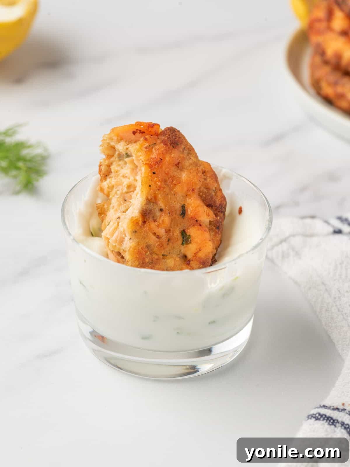 A close-up shot of a salmon patty being dipped into creamy tzatziki sauce, highlighting its crispy exterior and the delicious combination.