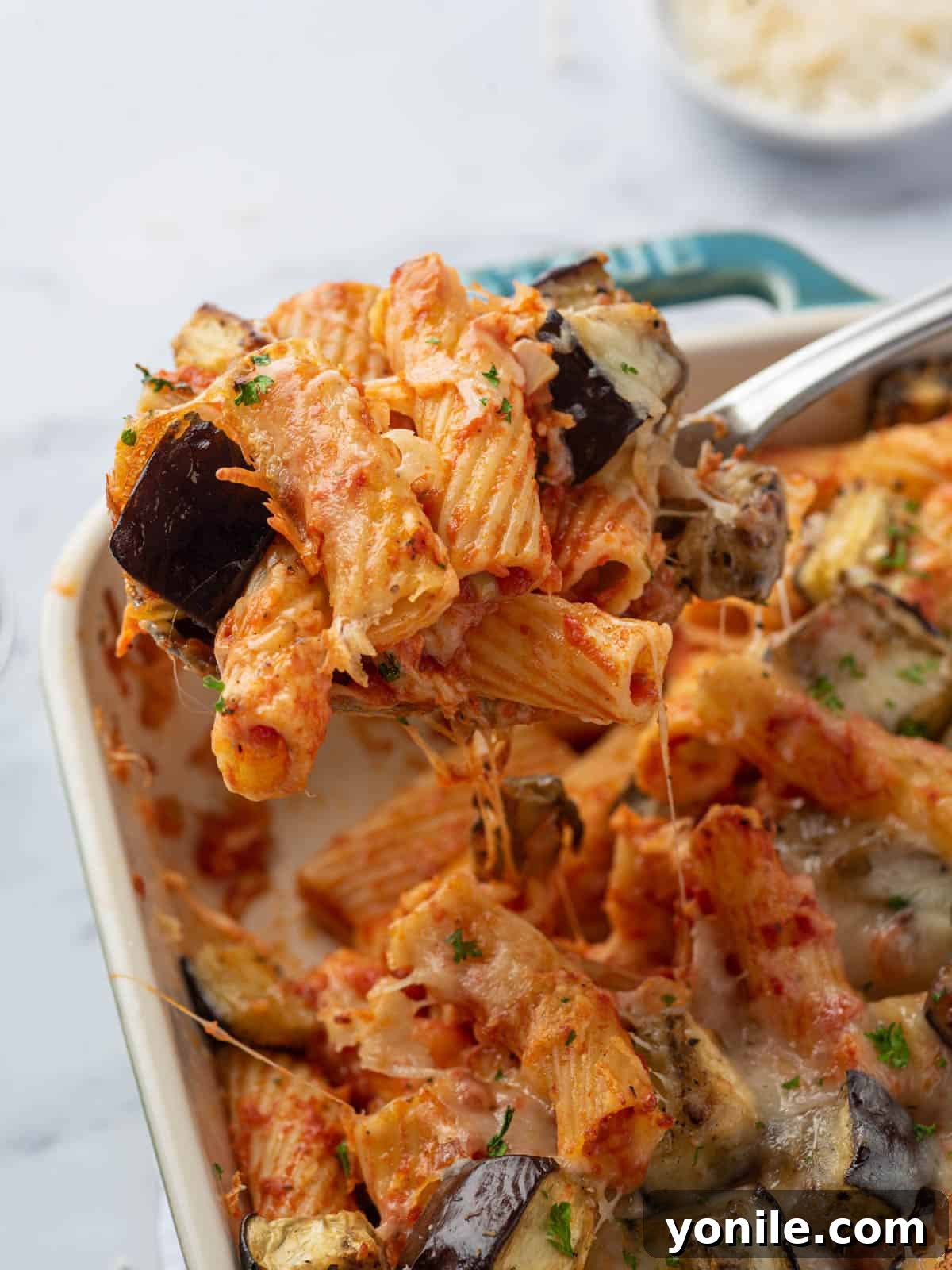 A serving of baked eggplant pasta on a white plate, with the full casserole dish blurred in the background.