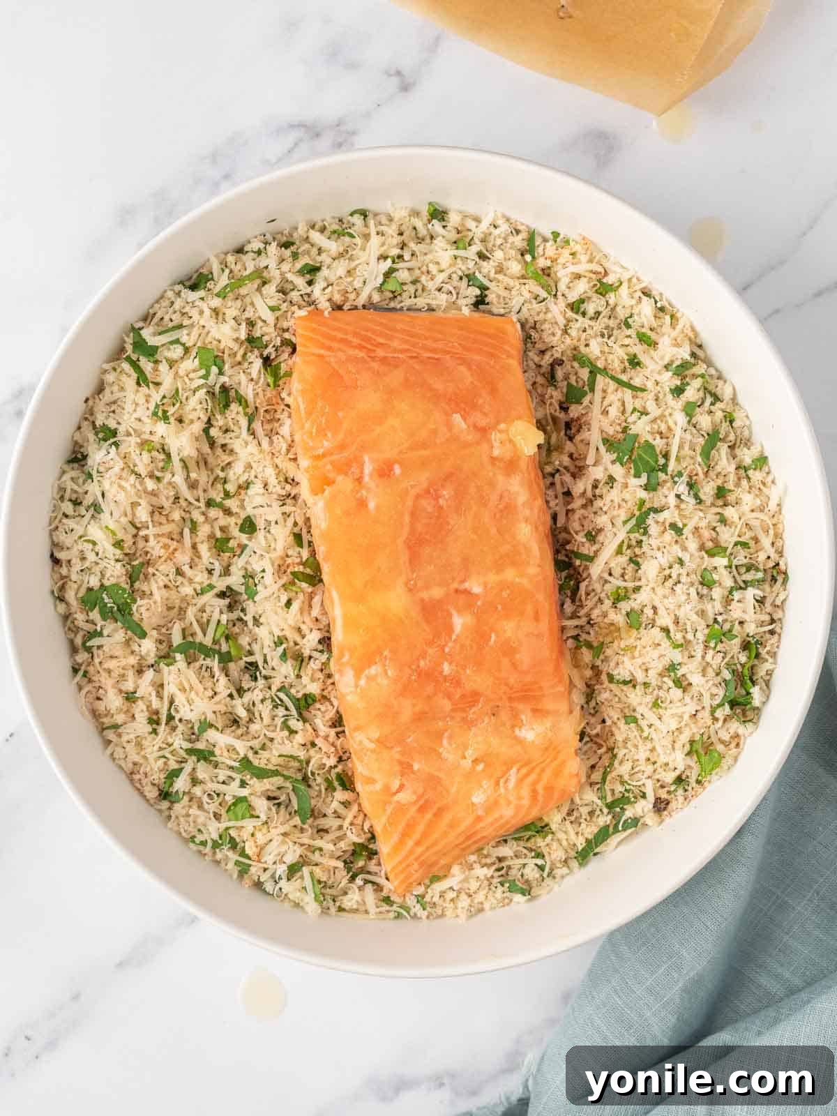 A close-up shows a salmon fillet being coated in a seasoned panko and parmesan mixture in a shallow bowl, ensuring an even, crisp crust.