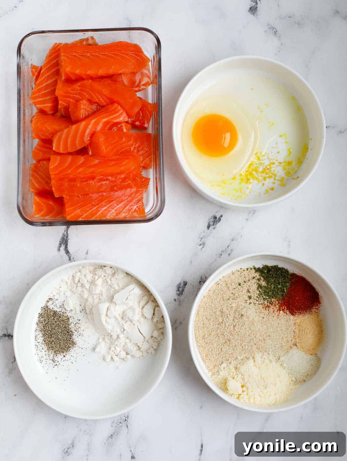 A selection of fresh ingredients laid out, including salmon fillets, various spices, flour, egg, and panko breadcrumbs, all essential for making crispy salmon fish fingers.