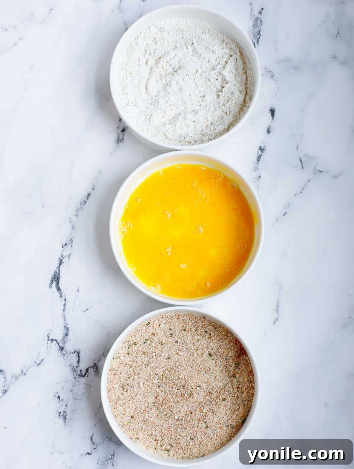 Three separate bowls containing the dredging station ingredients: flour mixture, whisked egg mixture, and panko breadcrumb mixture, all neatly arranged.