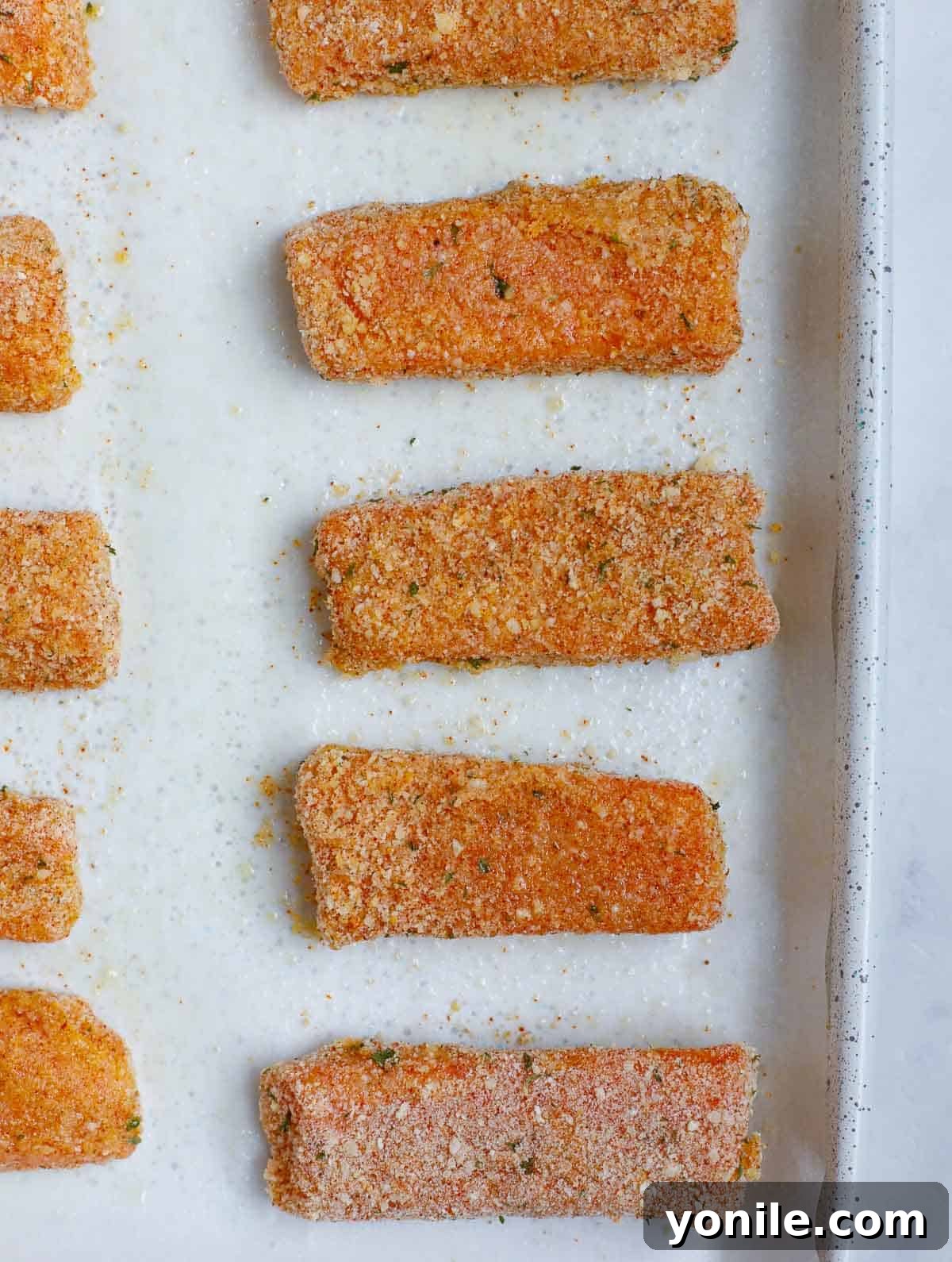 Numerous breaded salmon fish sticks neatly arranged in a single layer on a parchment-lined baking tray, ready to be placed in the oven.