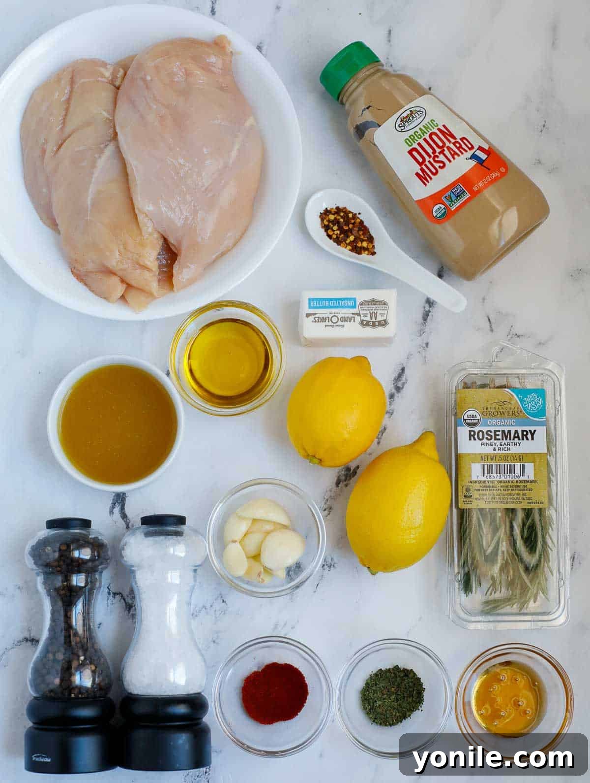 Fresh ingredients laid out on a cutting board, including chicken breasts, lemons, rosemary sprigs, garlic, and butter, ready for cooking.