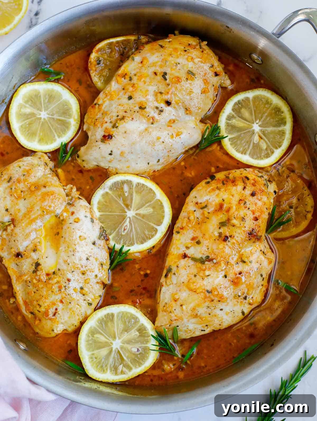 An overhead view of a hot skillet of baked rosemary chicken, garnished with fresh rosemary sprigs and lemon slices.