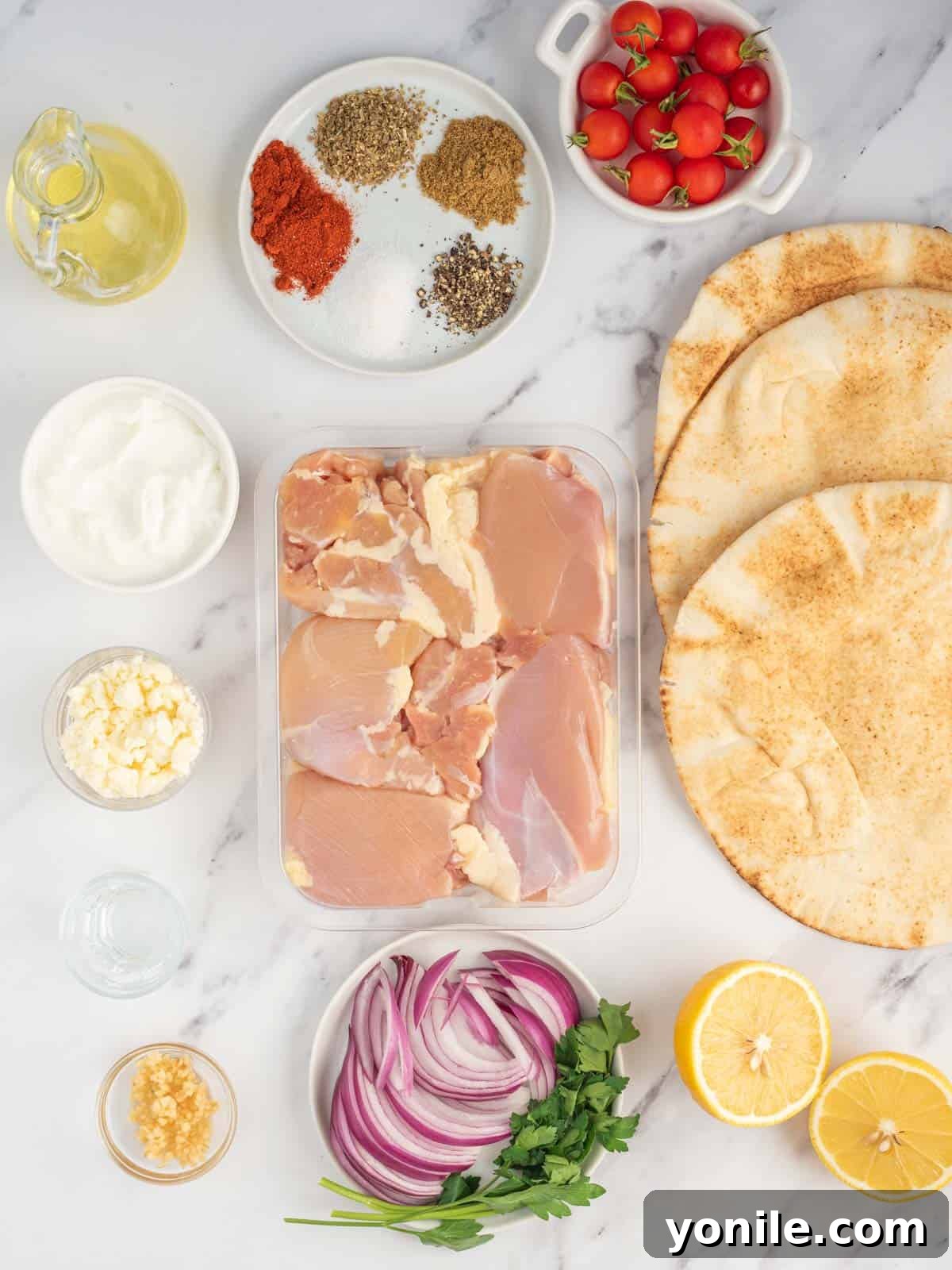 Authentic Homemade Greek Chicken Gyros 3 A collection of fresh ingredients laid out, including Greek yogurt, lemon, garlic, herbs, chicken, pita bread, tomatoes, red onion, and feta cheese, for making homemade Greek chicken gyros.