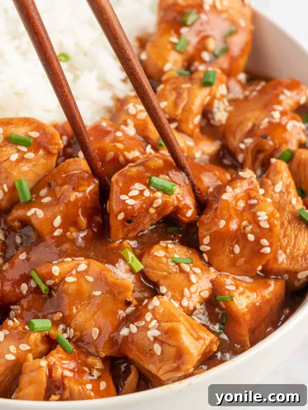 Chopsticks pick up a piece of sesame chicken from a bowl.
