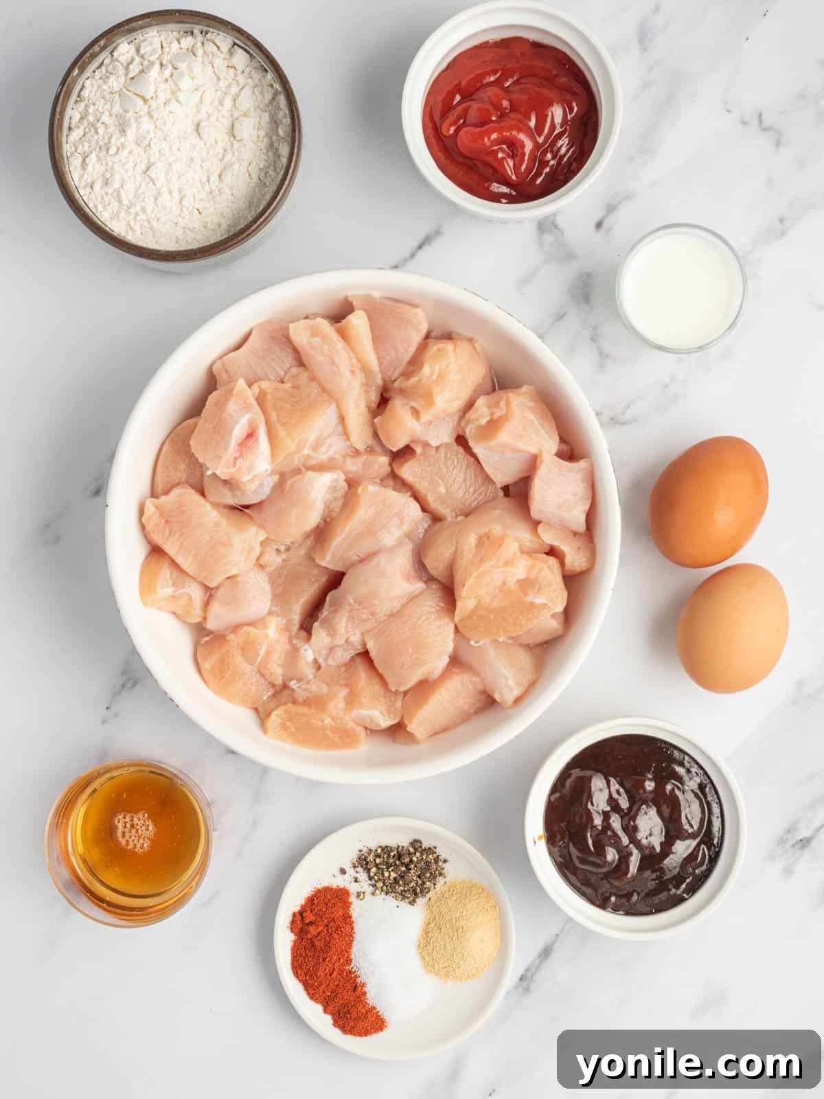 Collection of fresh ingredients for baked BBQ chicken bites, including chicken, seasonings, flour, eggs, milk, BBQ sauce, ketchup, and honey.