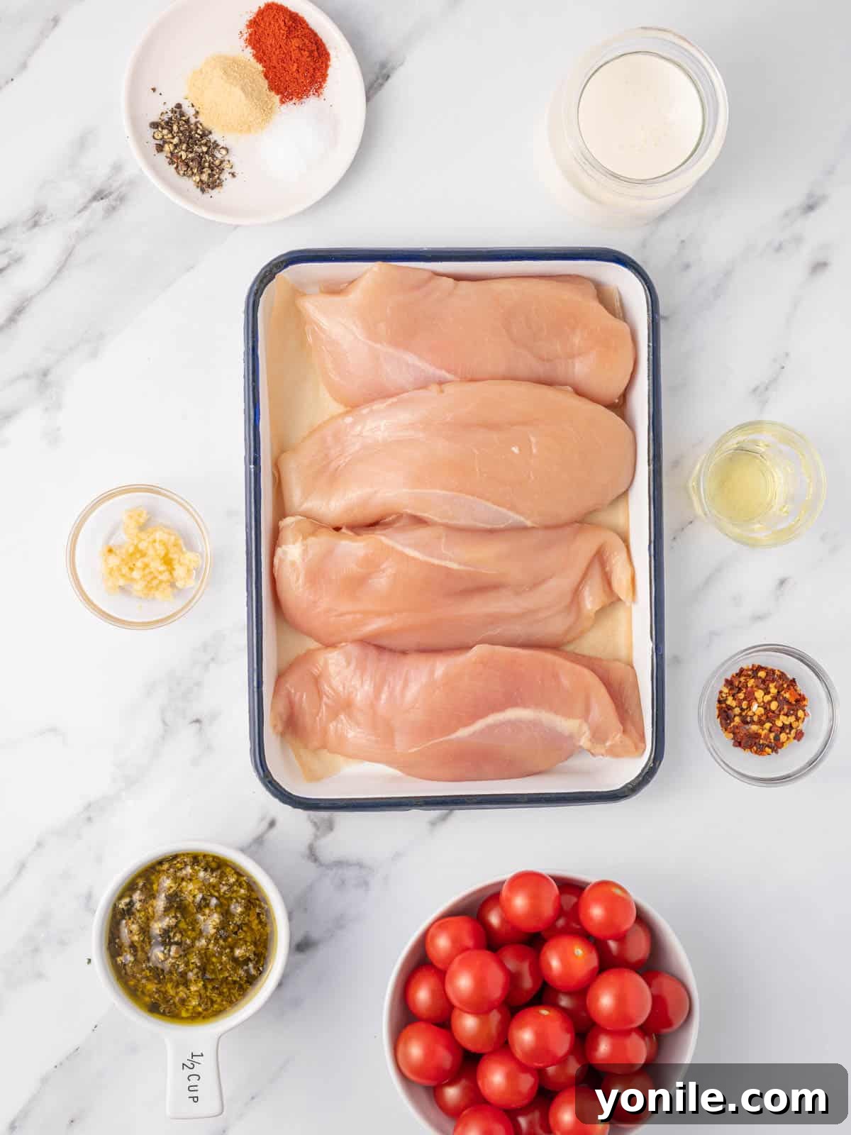 A flat lay photograph showcasing all the fresh ingredients needed for creamy pesto chicken, including chicken breasts, cherry tomatoes, basil pesto, heavy cream, garlic, and various seasonings.