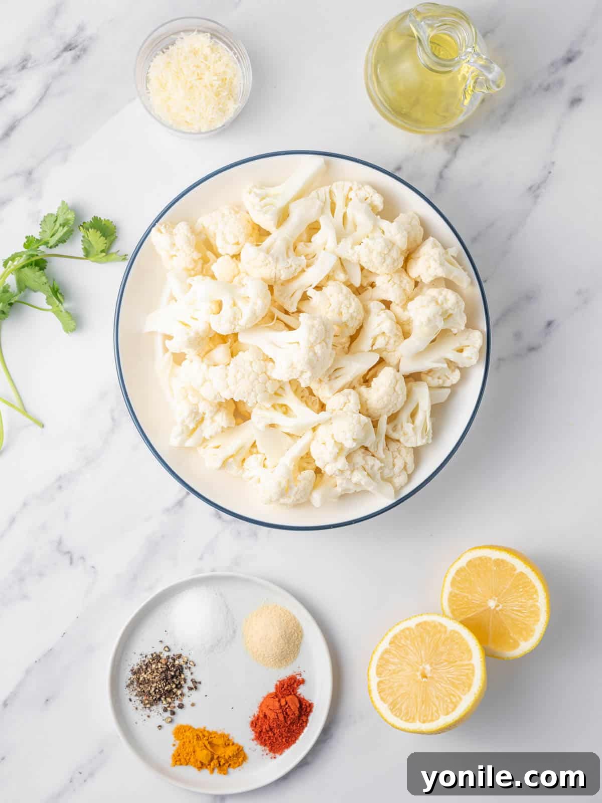A flat lay photograph showcasing all the fresh ingredients needed for making air fryer cauliflower bites, including a head of cauliflower, olive oil, parmesan, cilantro, and various spices.