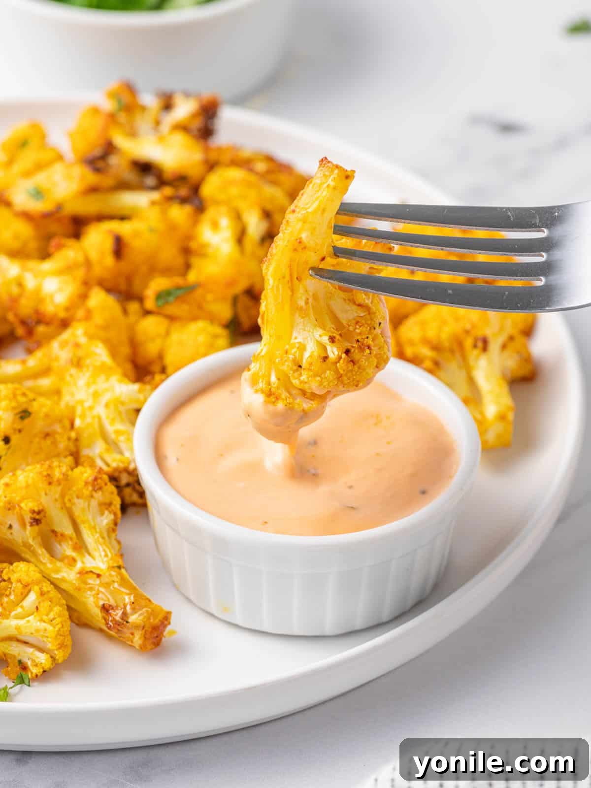 A fork piercing a single crispy air fryer cauliflower bite and dipping it into a creamy white sauce.