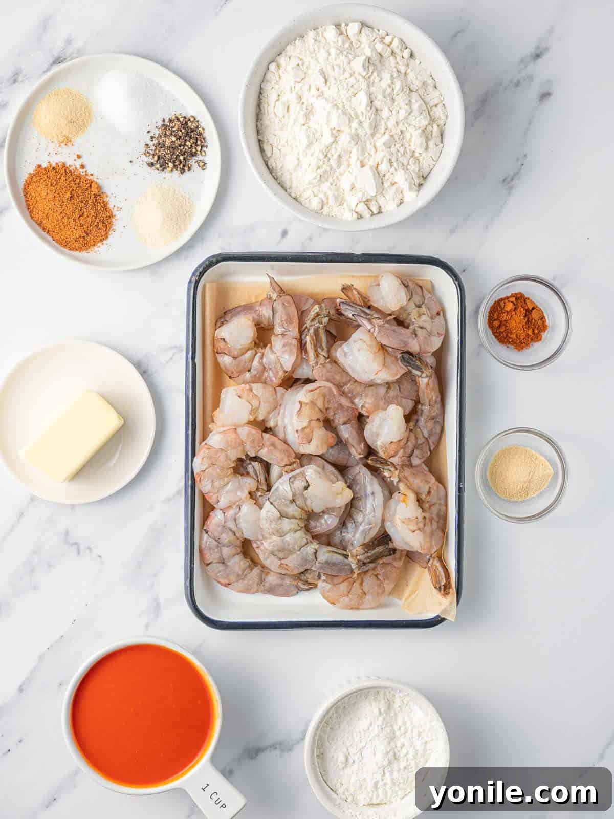 A selection of fresh ingredients laid out for making buffalo shrimp, including shrimp, flour, spices, hot sauce, and butter.