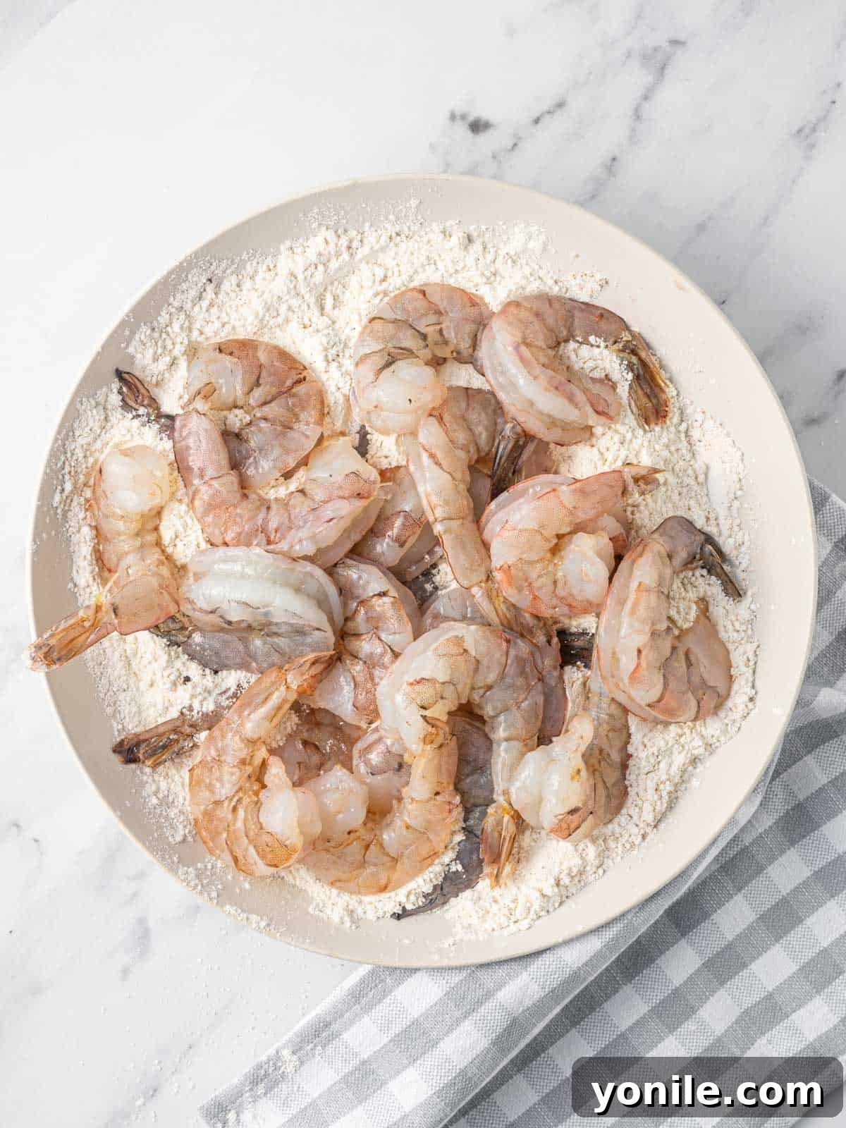 Hands coating raw shrimp thoroughly with a seasoned flour mixture in a bowl, preparing them for frying.