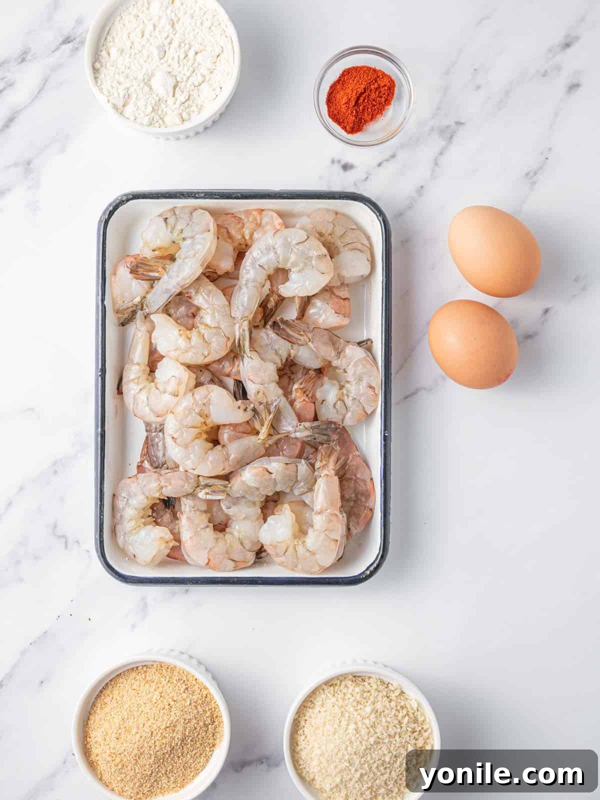 A flat lay photograph showcasing all the fresh ingredients required to prepare homemade butterfly shrimp, including raw shrimp, flour, eggs, breadcrumbs, and seasonings.