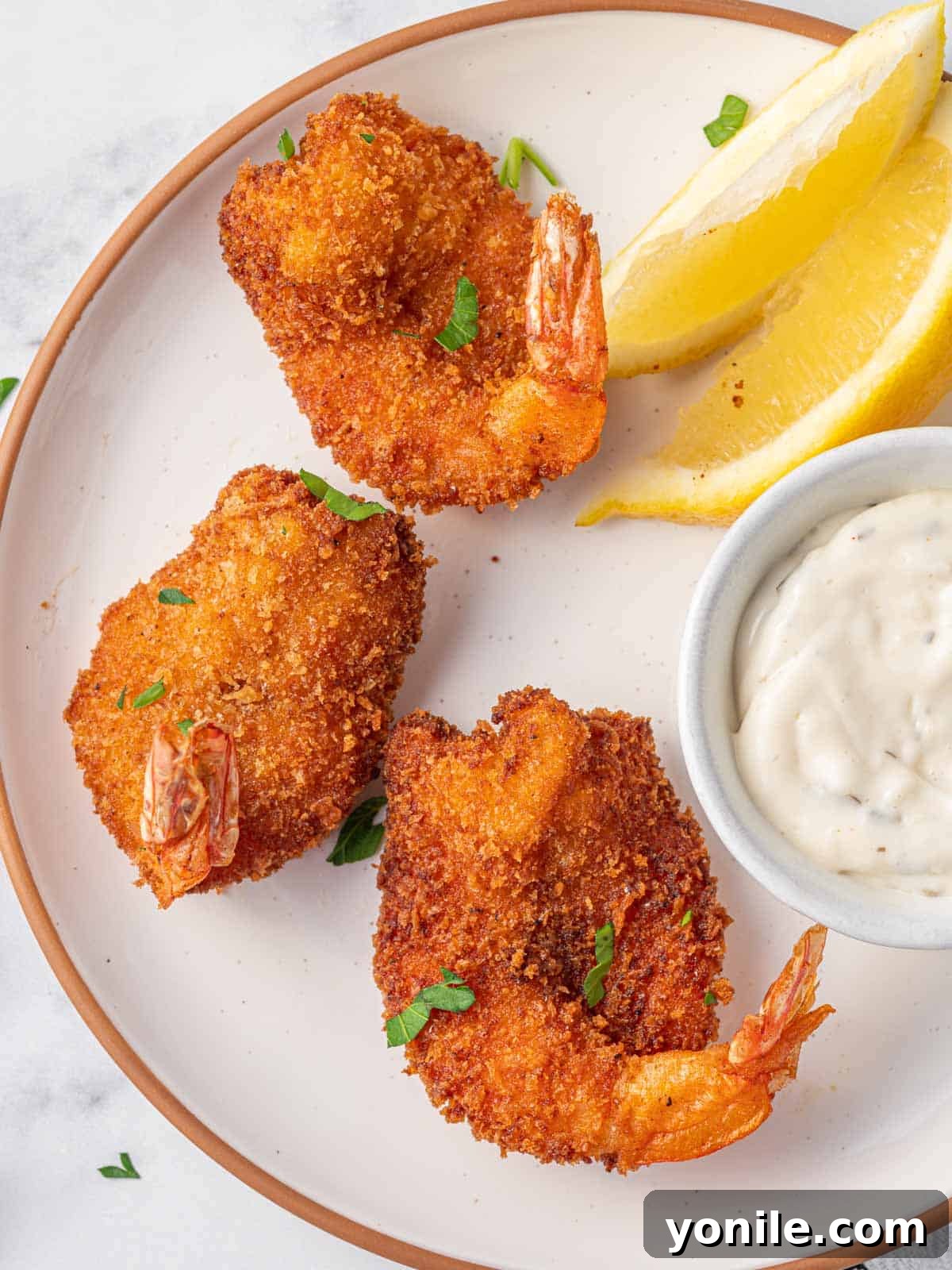Three golden-brown pan-fried butterfly shrimp elegantly presented on a white plate, accompanied by a small ramekin of creamy tartar sauce, ready for a gourmet experience.