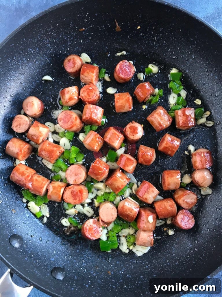 Close-up of golden-brown sausage pieces sizzling in a non-stick pan with sliced garlic and diced jalapeño.