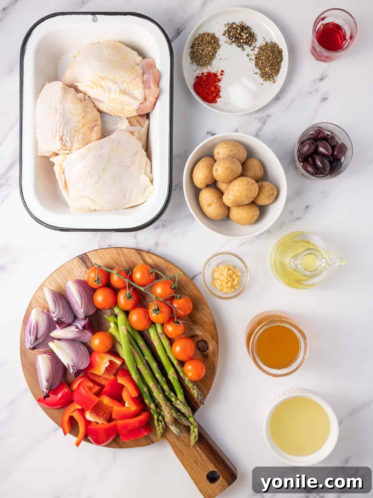 Fresh ingredients laid out for a Mediterranean baked chicken recipe, including raw chicken thighs, lemon, garlic, olive oil, red wine vinegar, various vegetables, and Kalamata olives.