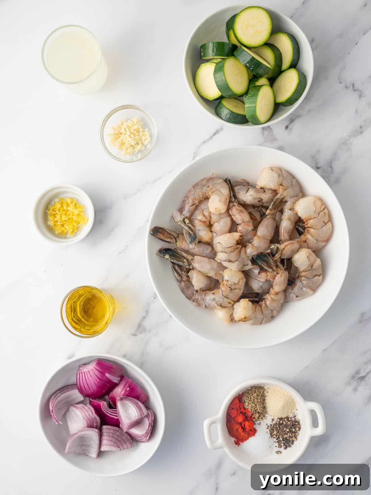 A collection of fresh ingredients laid out on a cutting board, including jumbo shrimp, sliced zucchini, cubed red onion, fresh lemons, a bottle of olive oil, and various spices, all ready for preparing marinated Greek shrimp skewers.