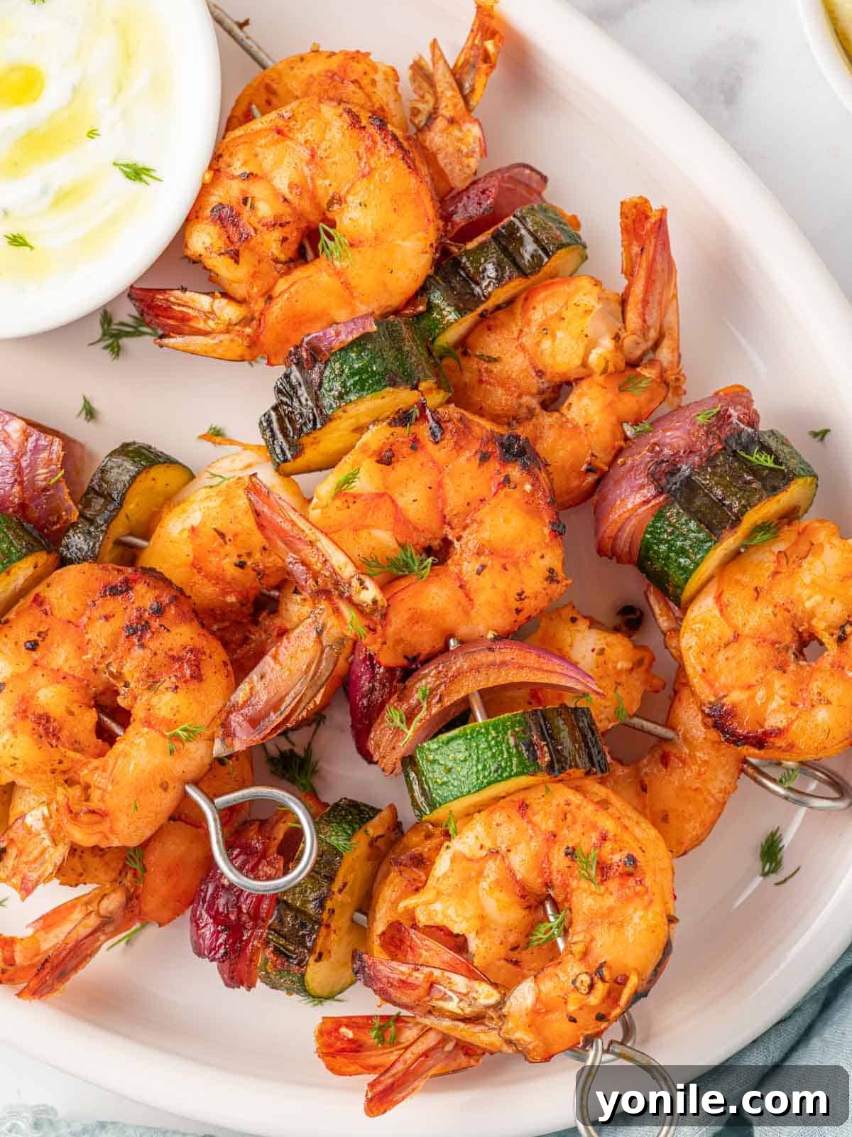 A close-up shot of several grilled shrimp skewers, featuring plump, pink shrimp interspersed with charred zucchini and red onion pieces, highlighting the delicious textures and vibrant colors.