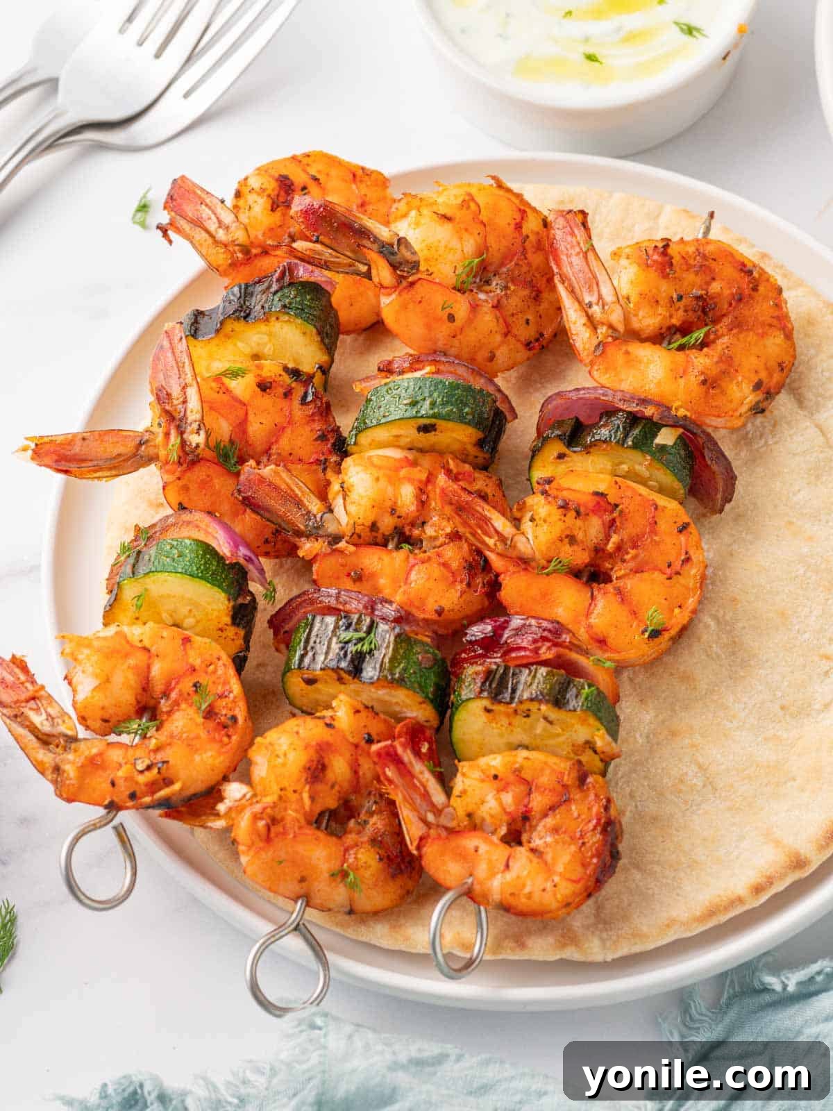 Three perfectly broiled Greek shrimp kabobs presented on a piece of pita bread, accompanied by a creamy dipping sauce in a small white bowl and garnished with fresh dill.