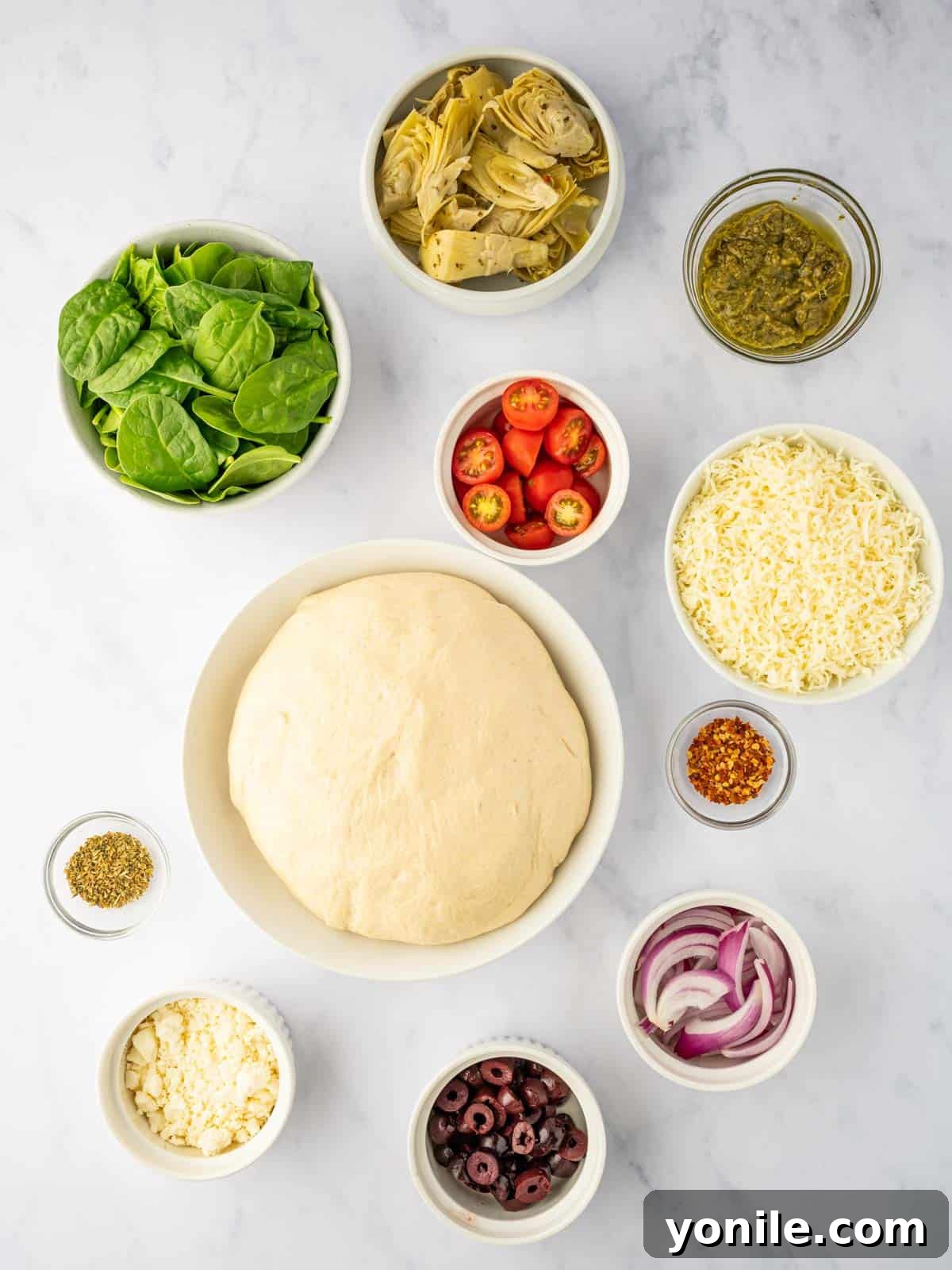 Homemade Mediterranean Pizza Feast 4 A vibrant selection of fresh Mediterranean ingredients including basil pesto, mozzarella, artichokes, spinach, tomatoes, olives, red onion, and feta cheese, ready for pizza topping.