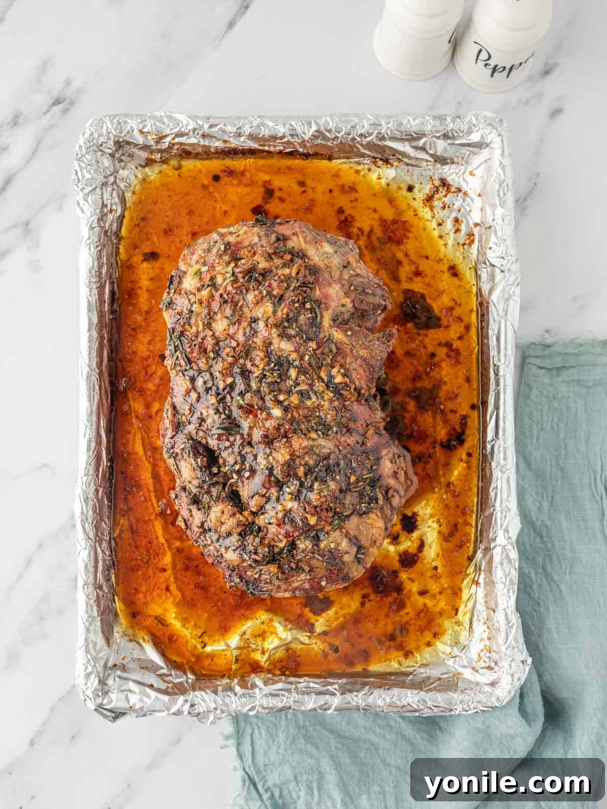 A perfectly roasted leg of lamb, gleaming with its own natural juices, resting gracefully in a foil-lined baking dish after its time in the oven.