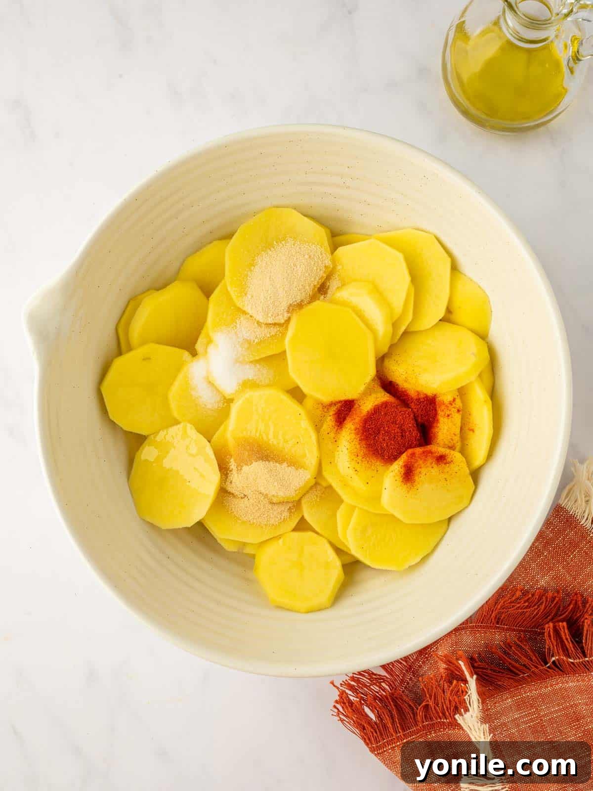 A hand sprinkling seasonings over potato slices in a mixing bowl.
