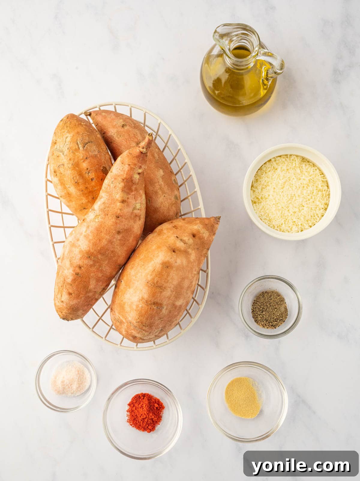 A carefully arranged flat lay of fresh sweet potatoes, a bottle of olive oil, a small dish of garlic powder, paprika, salt, pepper, and a bowl of grated parmesan cheese on a clean kitchen counter.