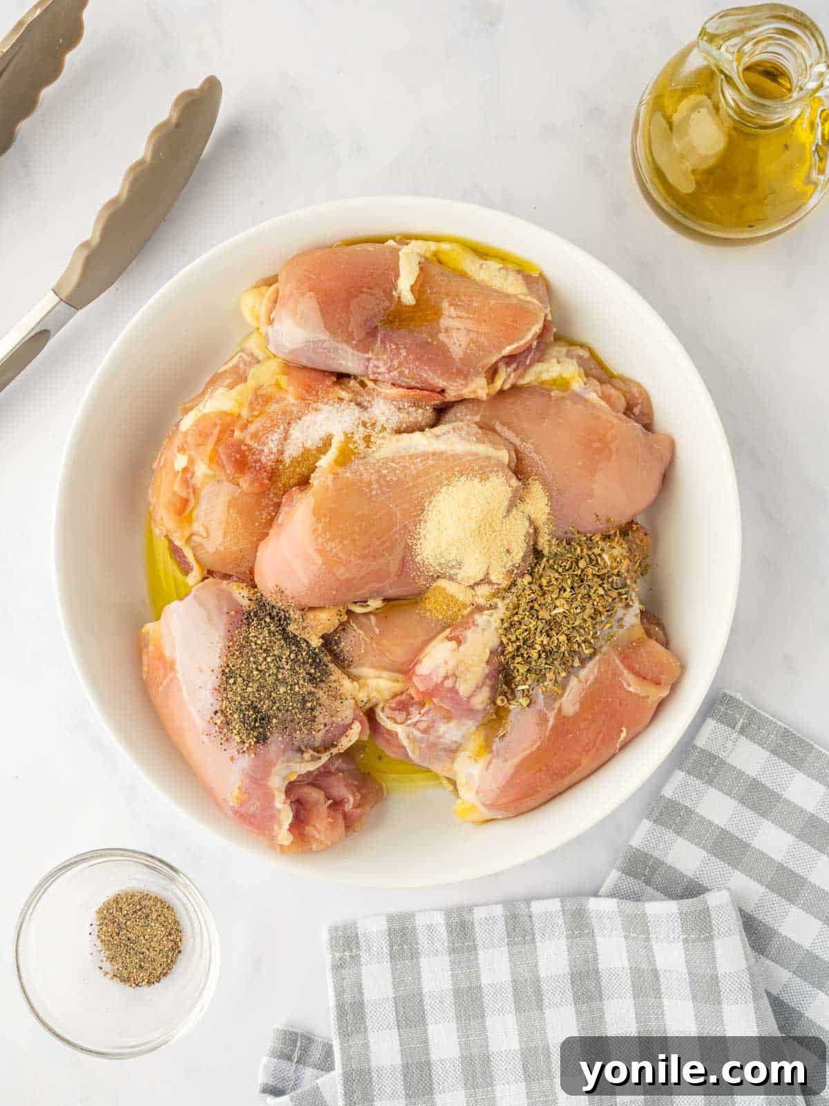 Raw chicken thighs are being tossed with olive oil and a blend of oregano, garlic powder, salt, and pepper in a mixing bowl.