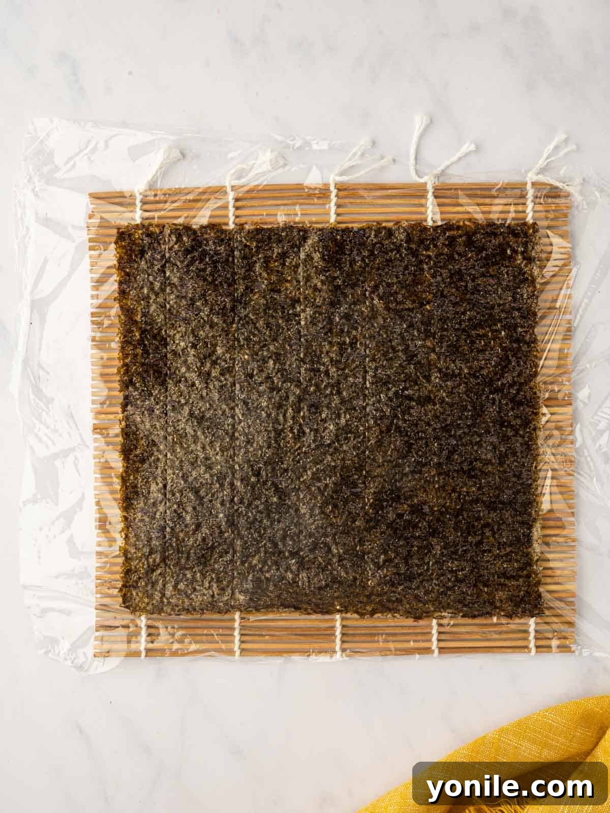 Dynamite Salmon Roll 8 A nori sheet is carefully placed over plastic wrap on a bamboo rolling mat, ready for sushi.