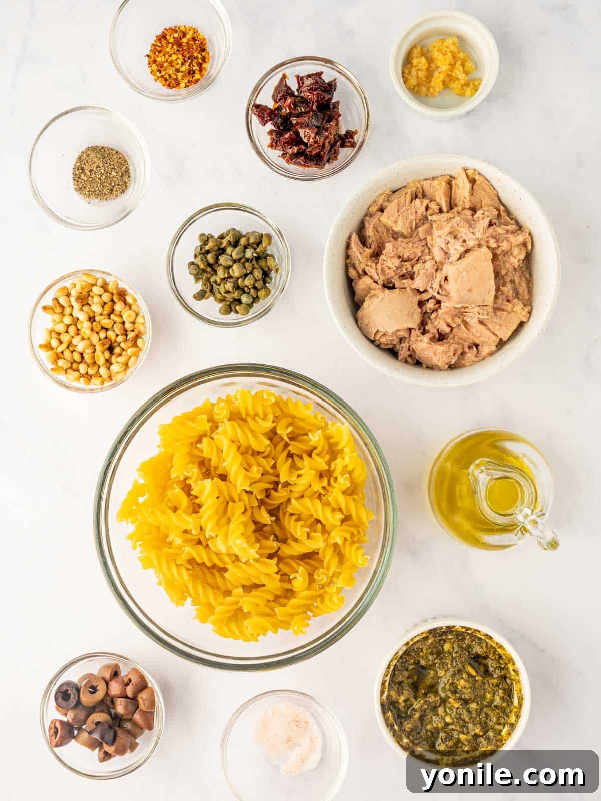 A spread of fresh ingredients for Tuna Pesto Pasta, including fusilli, canned tuna, sun-dried tomatoes, capers, garlic, and basil pesto.