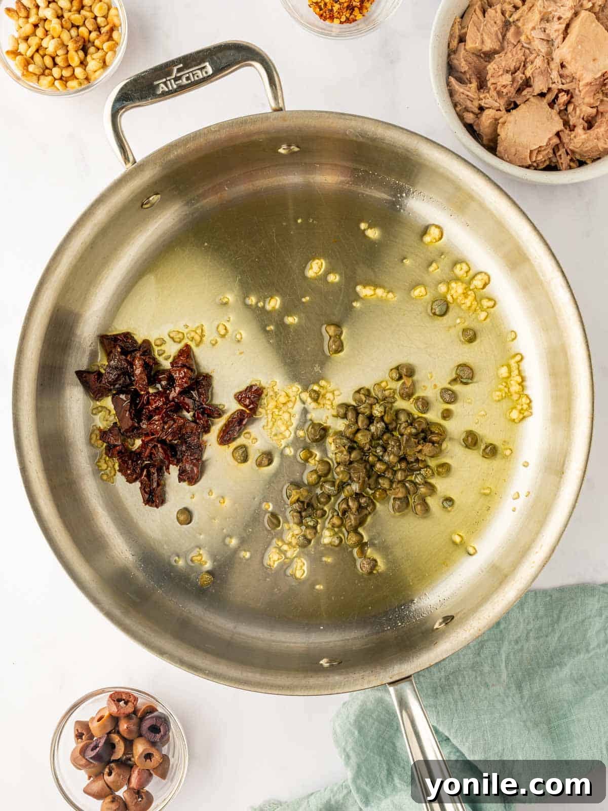 Fragrant capers, sun-dried tomatoes, and minced garlic are gently sautéed in olive oil in a skillet, creating an aromatic base for the pasta.