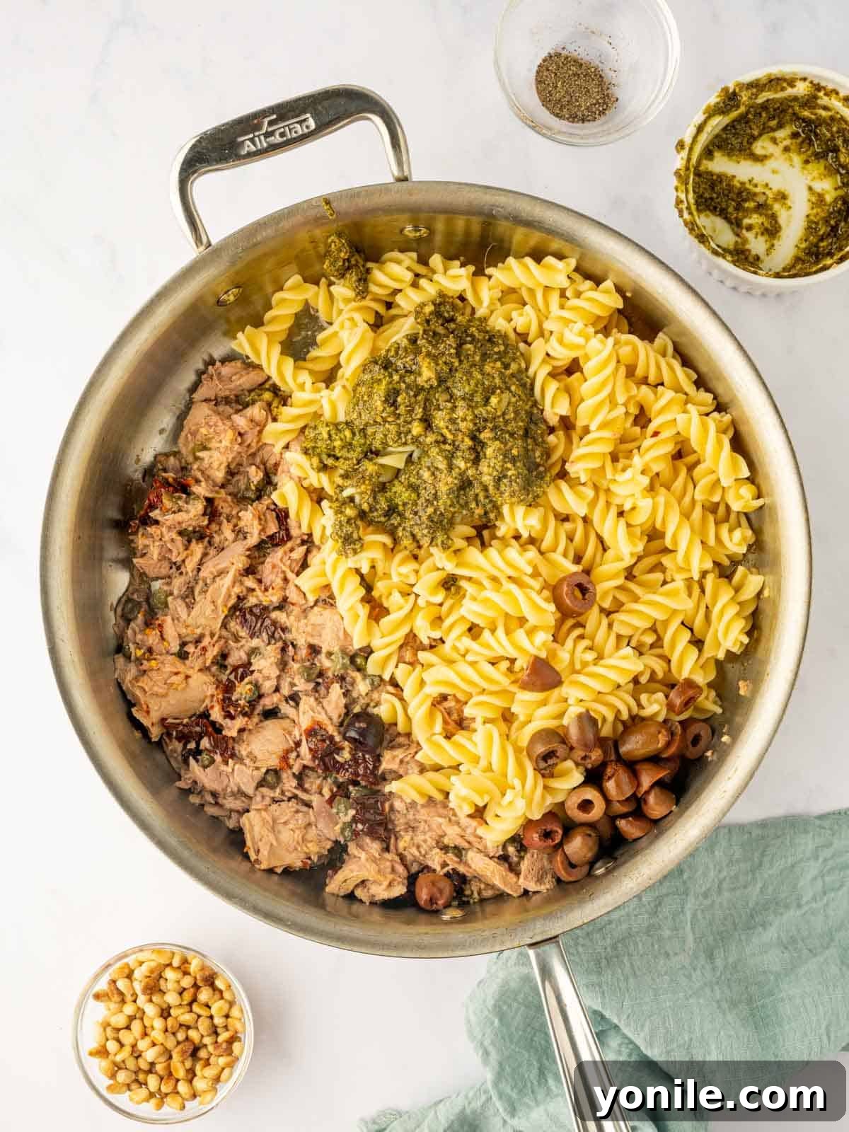 Cooked pasta, Kalamata olives, and vibrant basil pesto are artfully combined with the tuna, tomatoes, and capers in a large skillet, creating a harmonious mix.