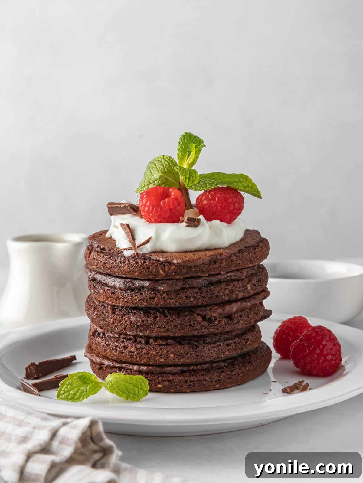 A tall stack of fluffy chocolate protein pancakes garnished with berries and a drizzle of syrup on a pristine white plate, promising a decadent yet healthy breakfast experience.
