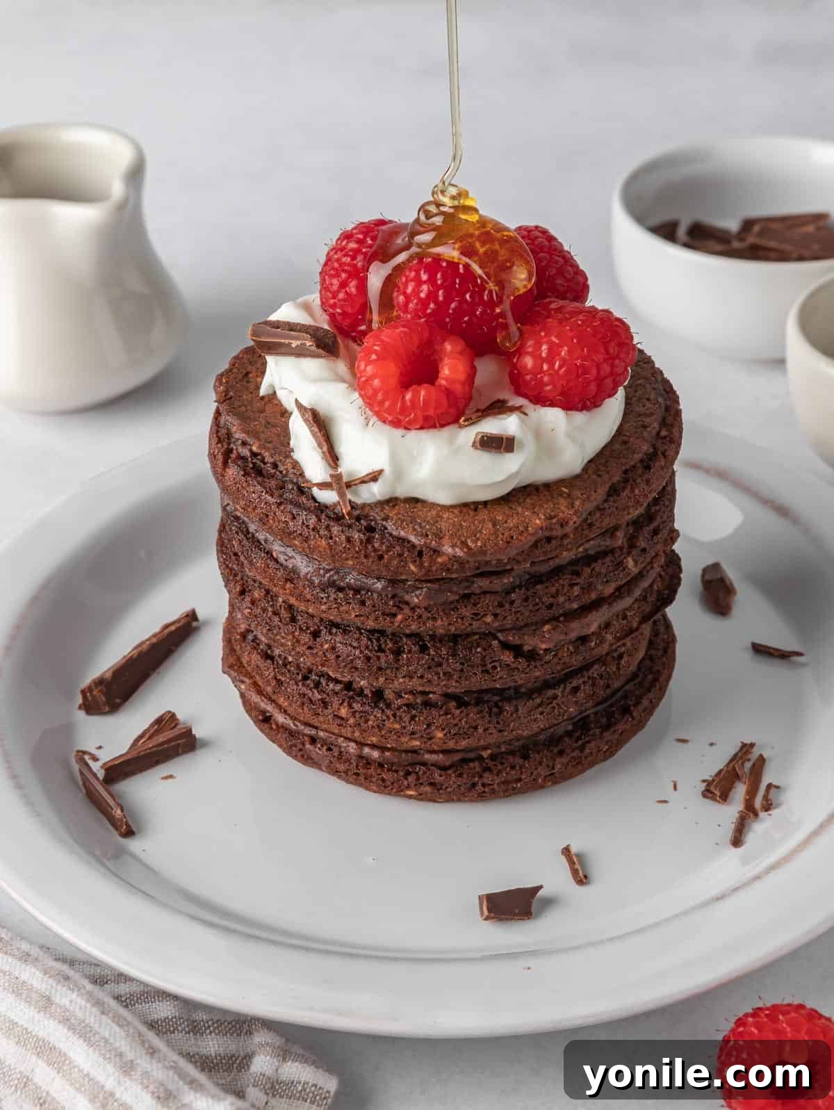 A stack of freshly cooked chocolate protein pancakes, beautifully topped with creamy whipped cream and vibrant fresh raspberries, ready to be served.
